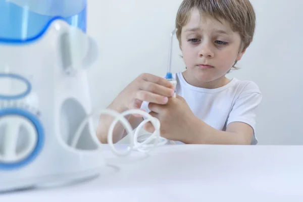Un niño de 5 años se sienta en casa en una mesa blanca con una camiseta blanca y se lava los dientes con un irrigador oral. el niño sonríe y sostiene el mango del irrigador . Fotos de stock