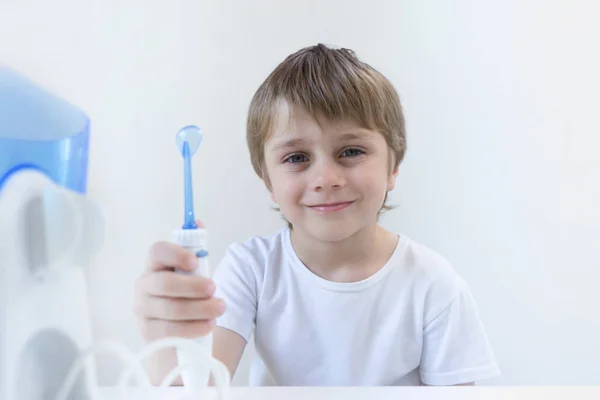 Un niño de 5 años se sienta en casa en una mesa blanca con una camiseta blanca y se lava los dientes con un irrigador oral. el niño sonríe y sostiene el mango del irrigador . Imagen de stock