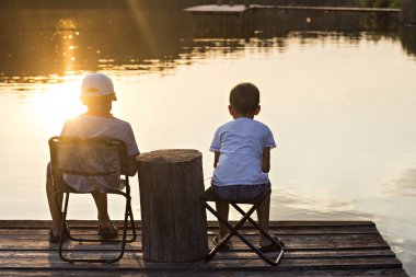 İki erkek nehir kıyısında oturmak ve günbatımında balık. Nehir kıyısında ellerinde sopalarla siting çocuklar