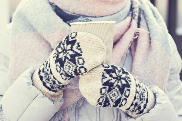 Female hands in white and black mittens holding steaming  cup of hot coffee cocoa, mulled wine or tea, outdoor. Winter time concept. Bask in the cold. Women\'s hands in mittens hold a craft paper glass