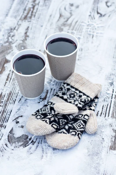 Two paper craft glass,  cup  with a hot drink, coffee, cocoa, mulled wine and white and black mittens on the table outdoor in the winter. Winter time concept. Bask in the cold.