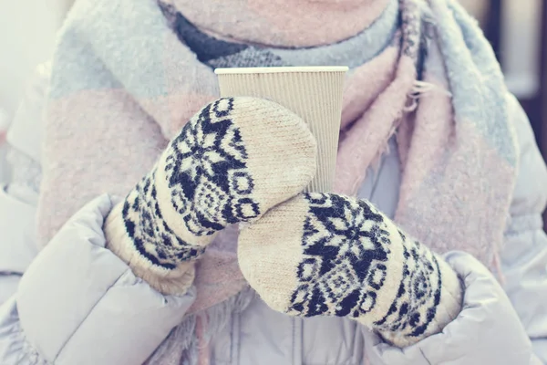 Female hands in white and black mittens holding steaming  cup of hot coffee cocoa, mulled wine or tea, outdoor. Winter time concept. Bask in the cold. Women\'s hands in mittens hold a craft paper glass