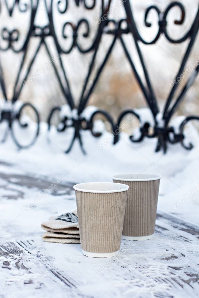 Two paper craft glass,  cup  with a hot drink, coffee, cocoa, mulled wine and white and black mittens on the table outdoor in the winter. Winter time concept. Bask in the cold. 
