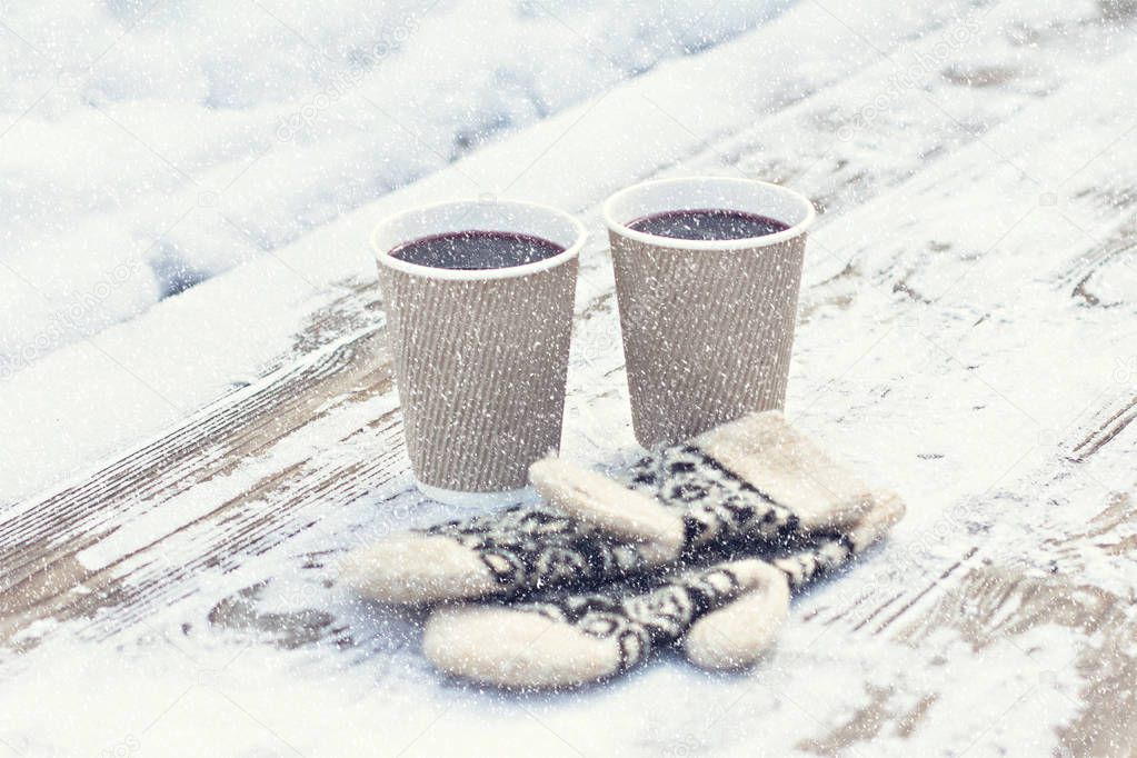 Two paper craft glass,  cup  with a hot drink, coffee, cocoa, mulled wine and white and black mittens on the table outdoor in the winter. Winter time concept. Bask in the cold. 