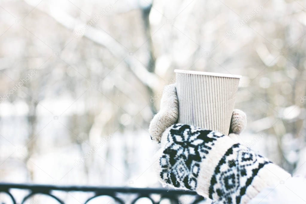 Female hands in white and black mittens holding steaming  cup of hot coffee cocoa, mulled wine or tea, outdoor. Winter time concept. Bask in the cold. Women's hands in mittens hold a craft paper glass