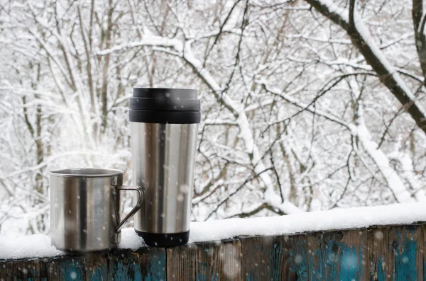 Cozy winter still life: Iron mug and thermo mug of hot coffee or tea on a snow covered terrace. Frosty winter day in forest. Christmas Holidays. Winter cozy background