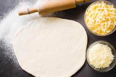 Siyah bir tahta üzerinde hamur, fırın un, rendelenmiş peynir ve haddeleme pin haddelenmiş.