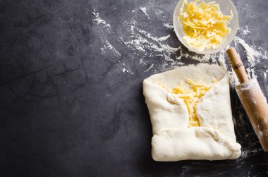 Siyah bir tahta üzerinde hamur, fırın un, rendelenmiş peynir ve haddeleme pin haddelenmiş.