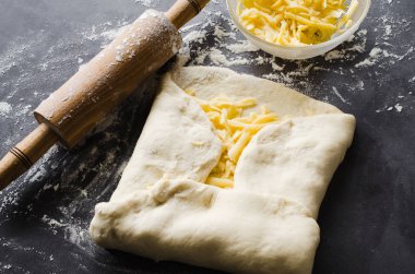 Siyah bir tahta üzerinde hamur, fırın un, rendelenmiş peynir ve haddeleme pin haddelenmiş.