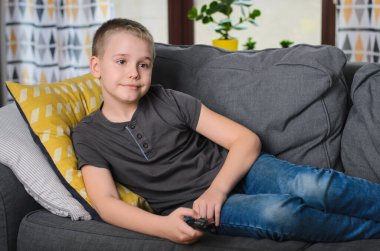 Genç çocuk kanepede uzanıp evde televizyon izliyor. Çocuk uzaktan kumandayı değiştiriyor. Seçici odak