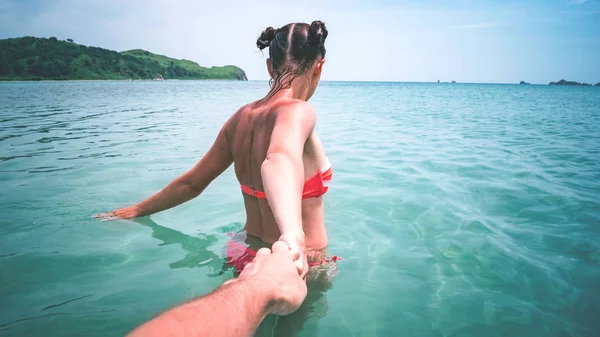 Summer vacation by the sea: a wet tanned girl in a red swimsuit enters the turquoise sea water and holds a man by the hand. Follow me.