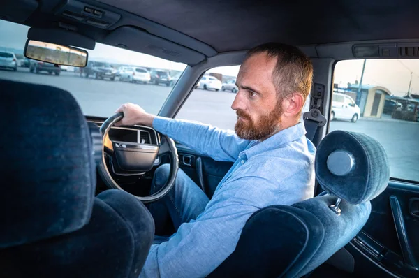 A man with a red beard in a blue shirt is driving a car in a parking lot. Blue car interior. Portrait.