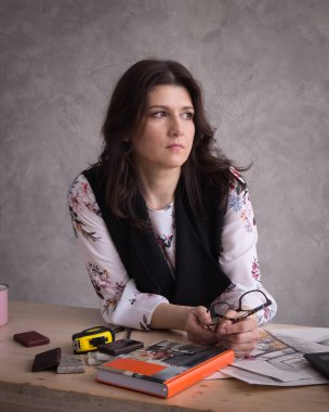 Yaratıcı meslekler: Bir yetişkin kadın tasarımcı iç tasarım hakkında düşünüyor ve gözlük onun elinde tutar. Masaya ölçme bant ve malzeme örnekleri bir dizüstü bilgisayardır. Portre.