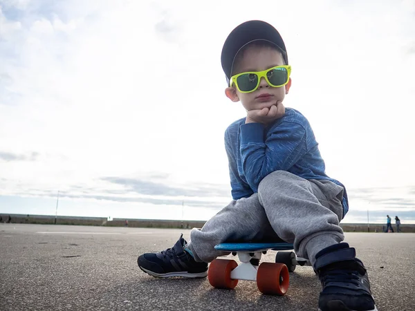 Straat sport voor kinderen: een jongetje in een pet en zonnebril ging zitten op een blauw bord cent om te ontspannen. — Stockfoto