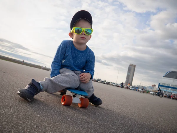Straat sport voor kinderen: een jongetje in een pet en zonnebril ging zitten op een blauw bord cent om te ontspannen. — Stockfoto