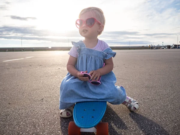 Lifstyle schieten: een gelukkig meisje in roze zonnebril en een blauwe jurk zit op een skate-board. — Stockfoto