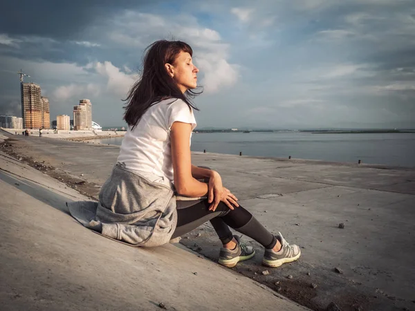 スポーツ ライフ スタイル 少女は市内の堤防に座っています ジョギングの後の息抜き 光の風の風髪 — ストック写真