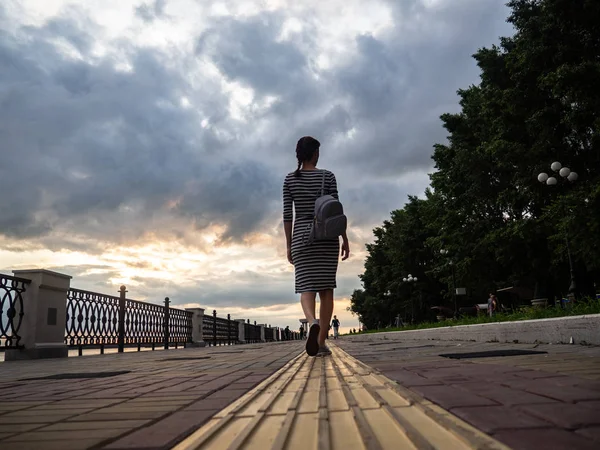 新鮮な空気の中を歩く 縞模様のガウンでバックパックを持つ少女が曇りの天候で堤防沿い散歩します 背面から見る — ストック写真