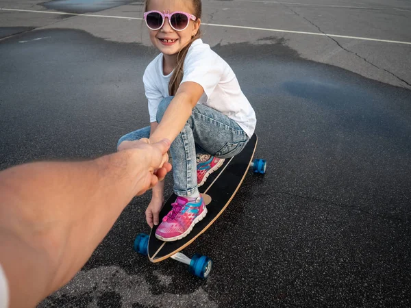 Dad helps my daughter to ride a log board. A girl in sunglasses sits on a logboard and holds her father\'s hand. Street portrait.