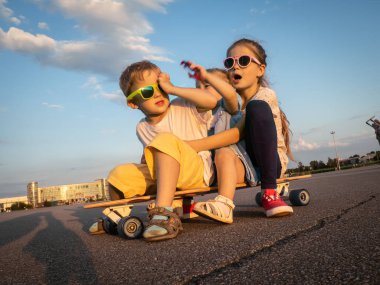 Sokak spor: güneş gözlüğü iki kız ve bir erkek birlikte büyük bir longboard üzerinde oturmak. Yaz solchechny çerçeve.