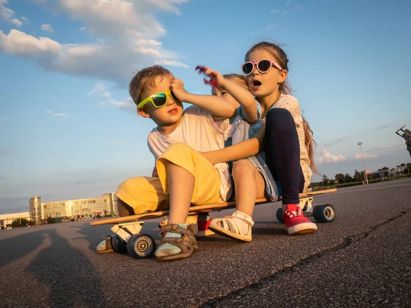 Straat sport: een jongen en twee meisjes in zonnebril samen zitten op een grote longboard. De zomer solchechny frame. — Stockfoto