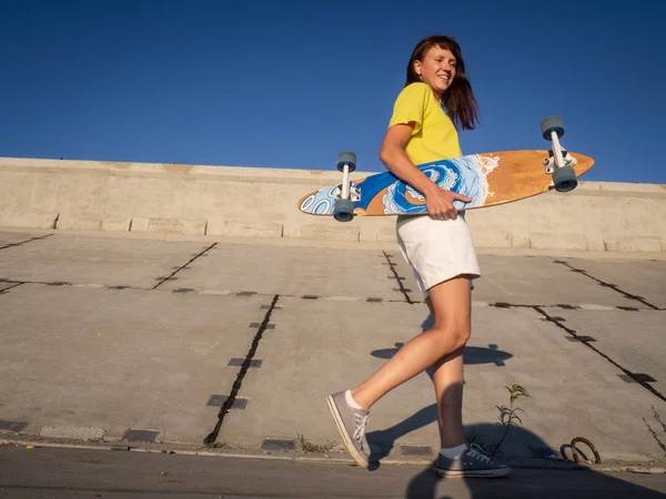 Straat sport: een vrolijk meisje in een geel T-shirt met een pintail longboard aan een betonnen kade loopt. Portret. — Stockfoto