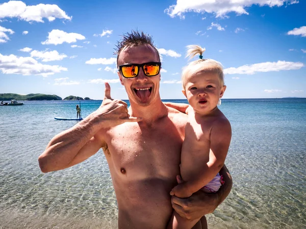 Vacaciones en la playa: Un tipo con gafas de sol en la orilla del mar sostiene a un niño en los brazos y muestra el símbolo de Aloha. Retrato . — Foto de Stock