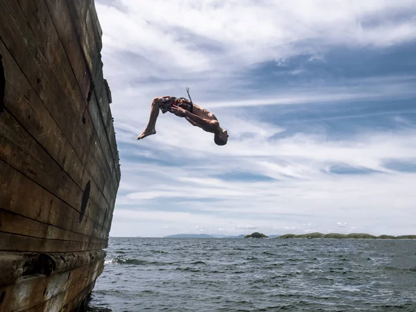 Cliff jumping: A young guy in shorts jumps into seawater from the side of an old ship.