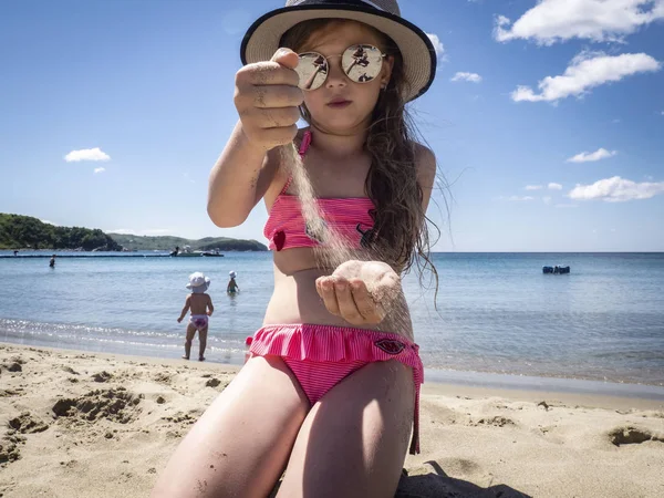 Vacations by the sea: a little girl in sunglasses and a hat pours her hands on the sand on a sea beach near the water.