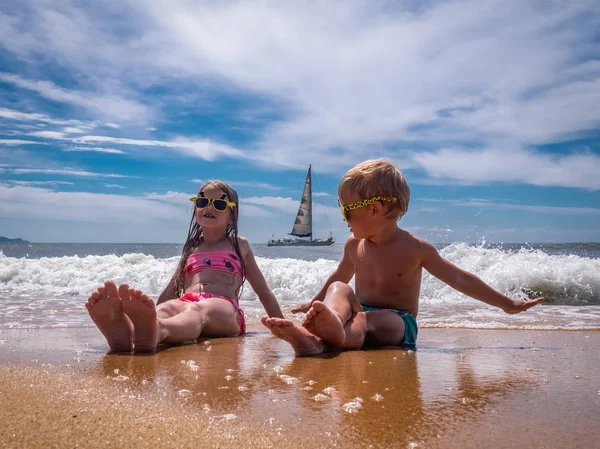 Children on the beach: A boy and a girl in funny sunglasses are sitting on the seashore in sea foam. In the background a sailboat sails. — Stock Photo, Image
