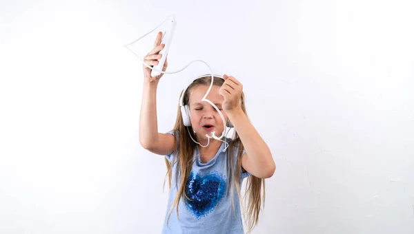 A girl in a blue t-shirt listens to music from a smartphone in large white headphones and sings along on a white background.