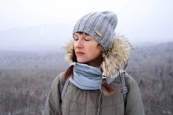 Hiking in the mountains: a girl in a knitted hat and with closed eyes breathes the fresh winter air. Portrait.