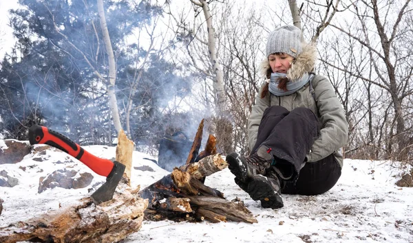 Hiking: A girl in a gray hat on a halt is warming herself by the fire in winter in the mountains.