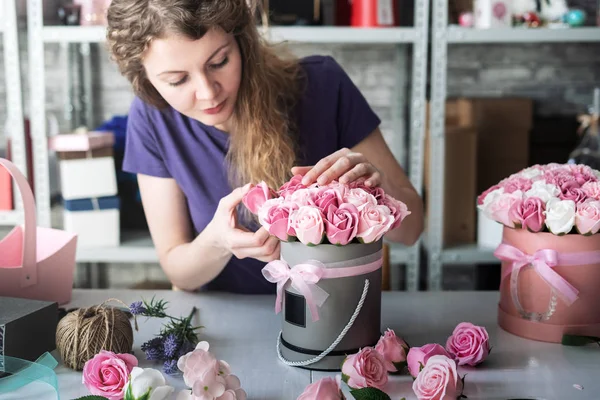 ร้านดอกไม้: สาวขายดอกไม้สะสมช่อดอกไม้ในกล่องกลมดอกกุหลาบสีชมพู ผมหยิกสีบลอนด์ หลังสีเทา . — ภาพถ่ายสต็อก