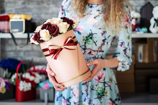 Flower Workshop: Florist girl is holding a small box with a bouquet of carnations and roses. Around on the shelves are many colorful bright bouquets.