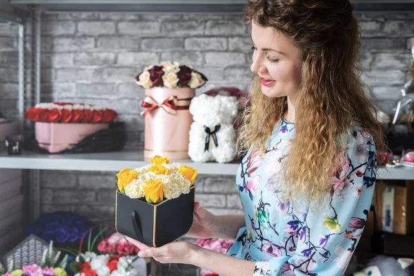 Flower Workshop: Florist girl is holding a small box with a bouquet of carnations and roses. Around on the shelves are many colorful bright bouquets.