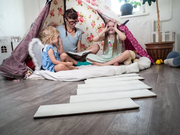 Tempo em família: mamãe e algumas das irmãs crianças brincam em casa em uma tenda caseira para crianças e mamãe lê um livro . — Fotografia de Stock