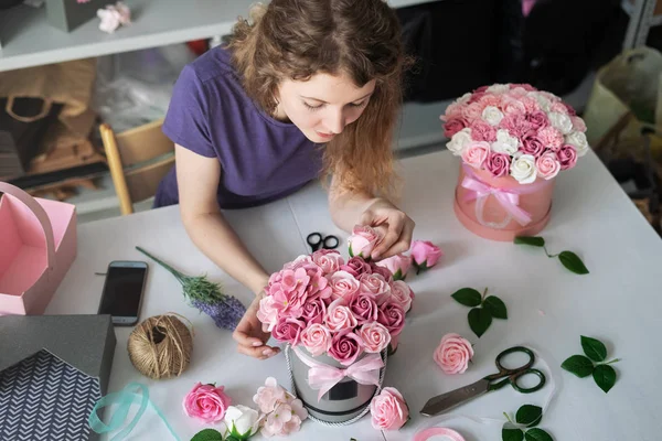 Floristería: una floristería recoge un ramo en una caja redonda de rosas rosadas. Cabello rubio rizado, fondo gris . — Foto de Stock