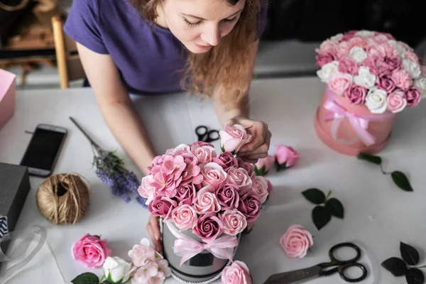 Floristería: una floristería recoge un ramo en una caja redonda de rosas rosadas. Cabello rubio rizado, fondo gris . — Foto de Stock