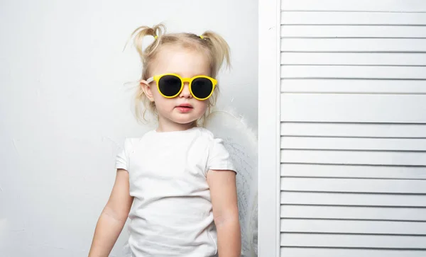 Moda infantil: una niña en gafas de sol amarillas posa seriamente contra una pared blanca, imitando a una modelo superior . — Foto de Stock