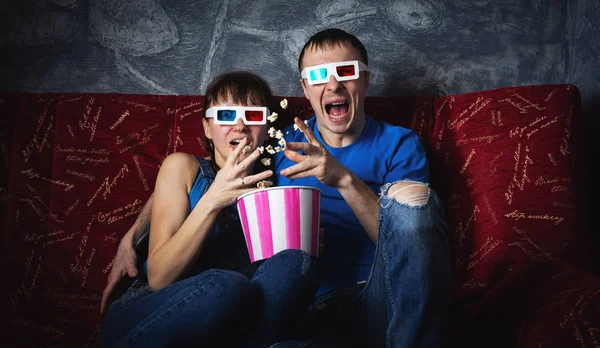 A couple of guy and girl watch a scary movie in 3D glasses and scream with fright. Popcorn.