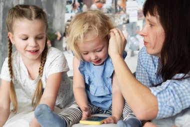 Anne ve iki kızı yatak ve anne düzgünleştirir kıvırcık saç üzerinde oturuyor. Genç bir smartphone ile oynuyor.