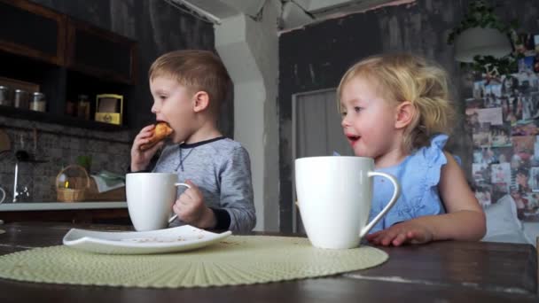 Un niño y una niña beben té de tazas grandes en la cocina familiar y comen croissants. Interior europeo . — Vídeos de Stock