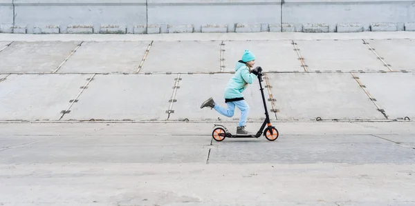 Activiteiten: Een school meisje met een blauwe jas een scooter rijden op een grijze betonnen promenade. — Stockfoto