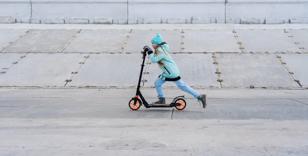 Activités : Une écolière avec une veste bleue chevauchant un scooter sur une promenade en béton gris . — Photo