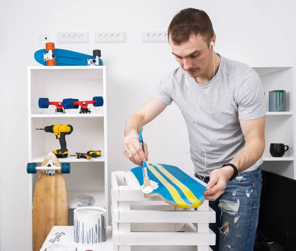 Um homem com uma camiseta cinza está pintando um skate em uma oficina e ouvindo música em fones de ouvido . — Fotografia de Stock