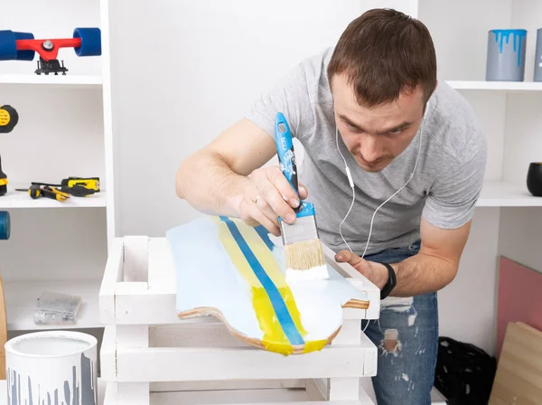 Um homem com uma camiseta cinza está pintando um skate em uma oficina e ouvindo música em fones de ouvido . — Fotografia de Stock