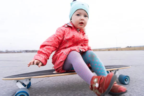 Een baby meisje in een blauwe hoed en roze jas is rollen op een Longboard in de stad. — Stockfoto