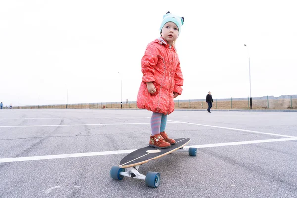 Une petite fille avec un chapeau bleu et une veste rose roule sur un longboard dans la ville . — Photo