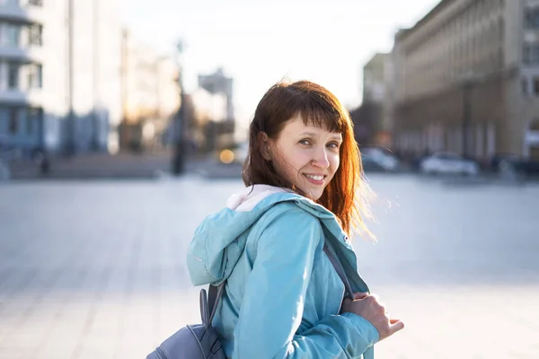 A menina em um blusão azul e com uma mochila caminha pela praça da cidade e olha para trás por cima de seu ombro. Fundo desfocado . — Fotografia de Stock